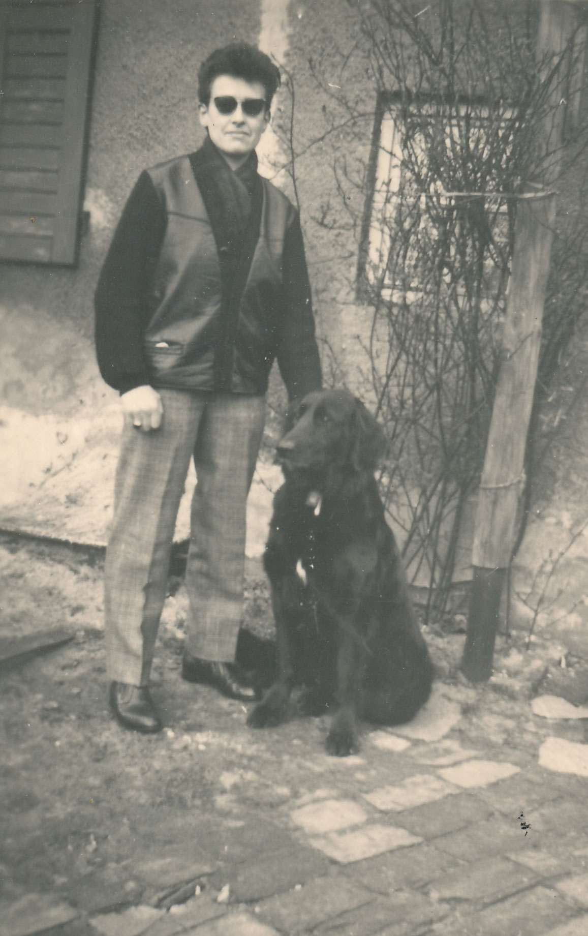 Tommy steht auf der Straße mit einem schwarzen Hund. Sie trägt eine Lederweste und eine Sonnenbrille.