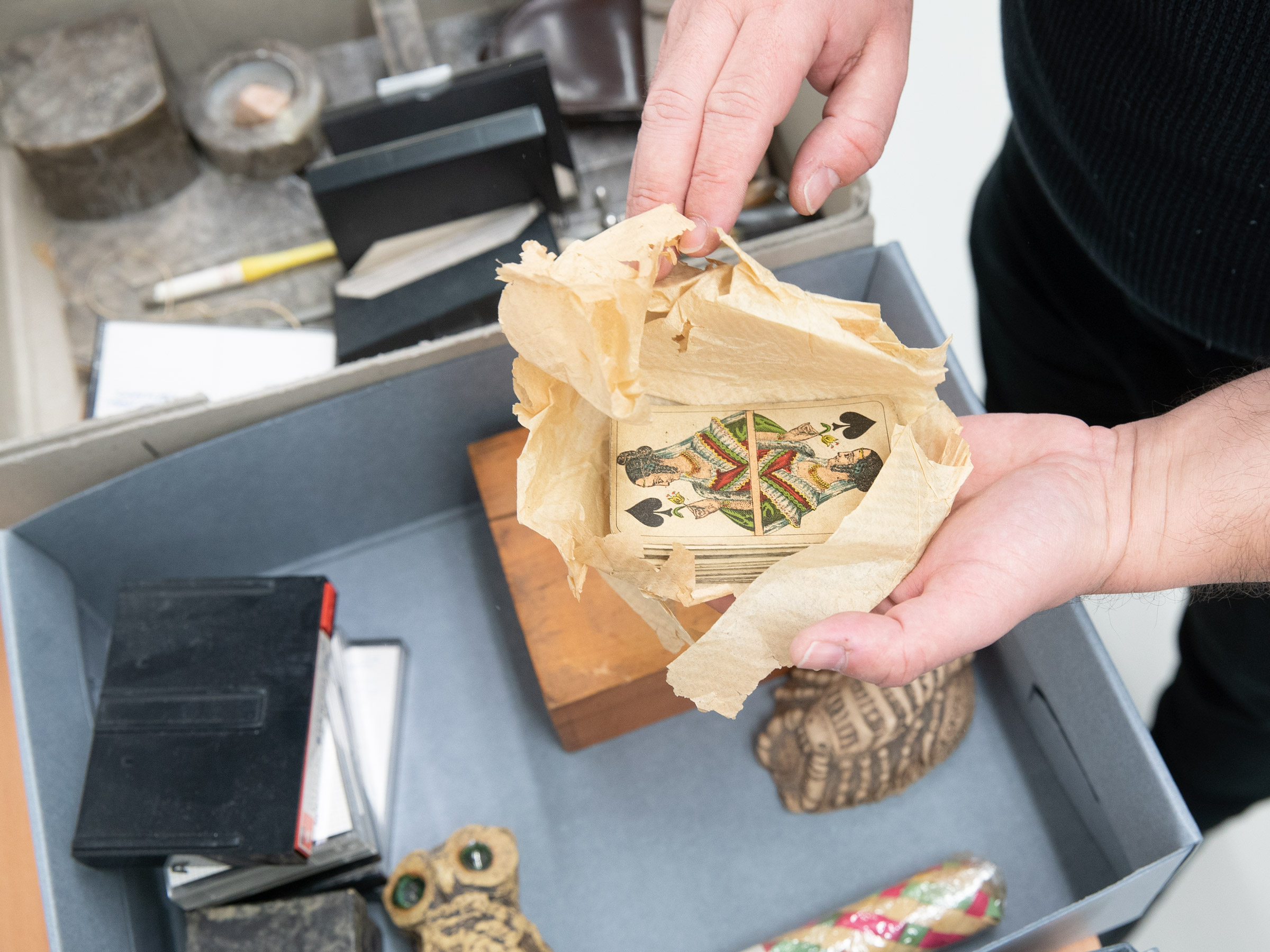 geöffnete Archivkiste mit diversen Objekten u. a. Kassetten, Holzschachtel und Figuren; Kartenspiel mit oben liegender Pik-Ass-Karte in den Händen eines Archivmitarbeiters