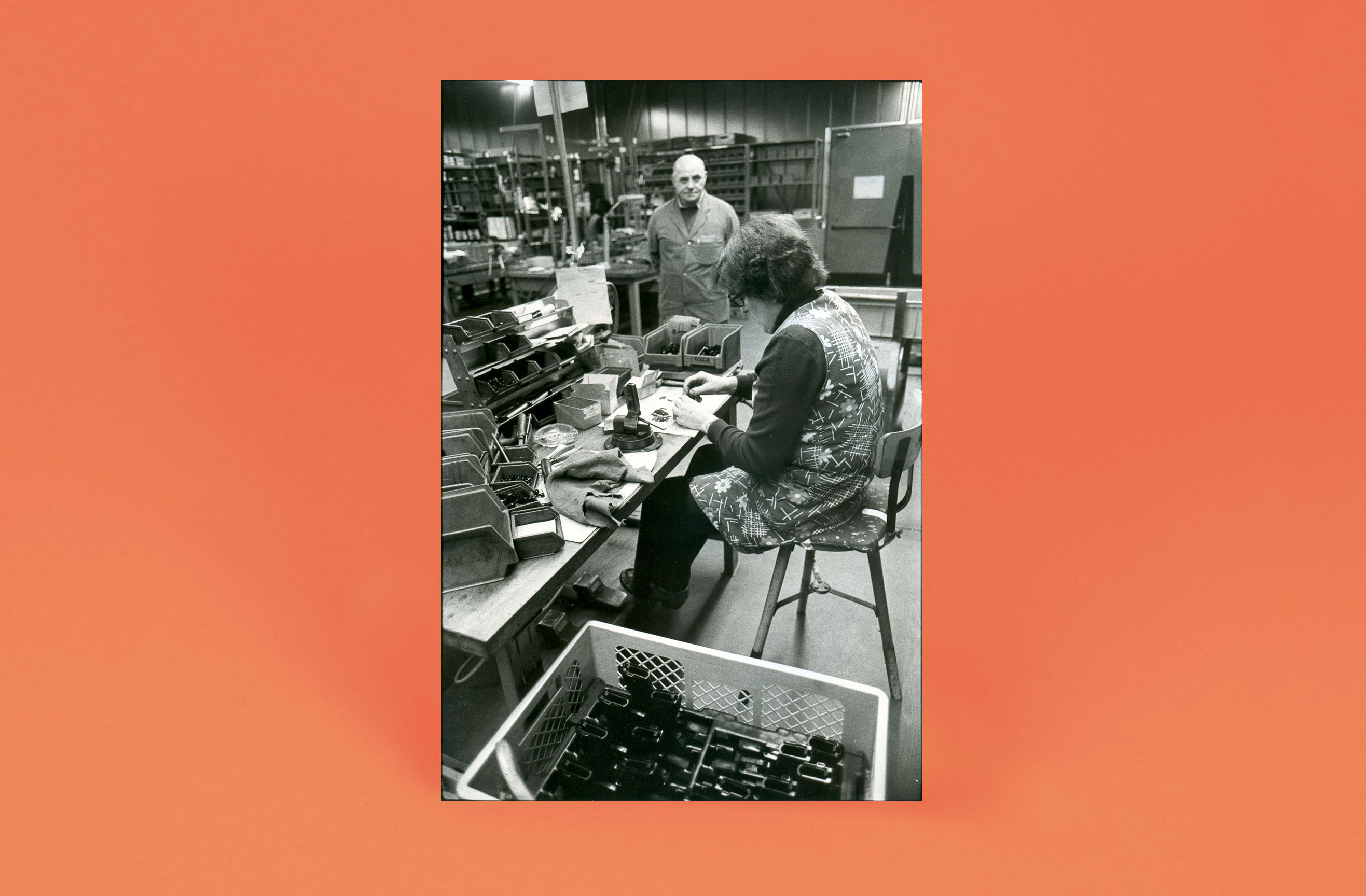 Frau sitzt in einer Fabrikhalle an einer Werkbank und setzt Metallteile von Pistolen mit der Hand zusammen; Mitarbeiter steht im Hintergrund