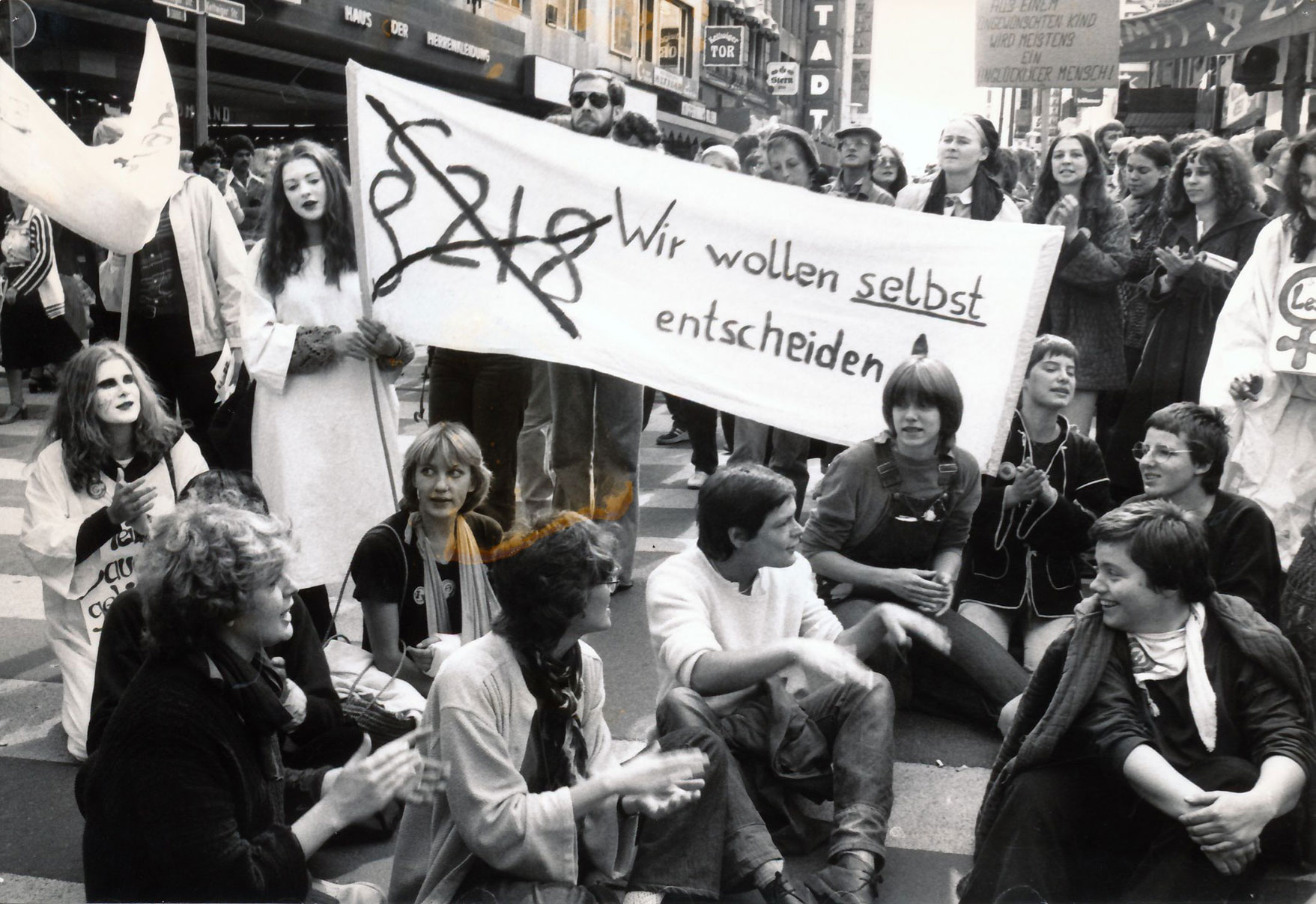 Demonstrantinnen sitzen und stehen auf der Straße, unter ihnen zwei junge Frauen in weißen Kitteln mit weiß geschminkten Gesicht, eine von ihnen hält ein Transparent mit dem durchgestrichenen Zeichen § 218 und der Aufschrift „Wir wollen selbst entscheiden!“; vorne Dagmar Brüssow im weißen Pullover auf dem Boden sitzend, dahinter in Christine Landgraf in Latzhose