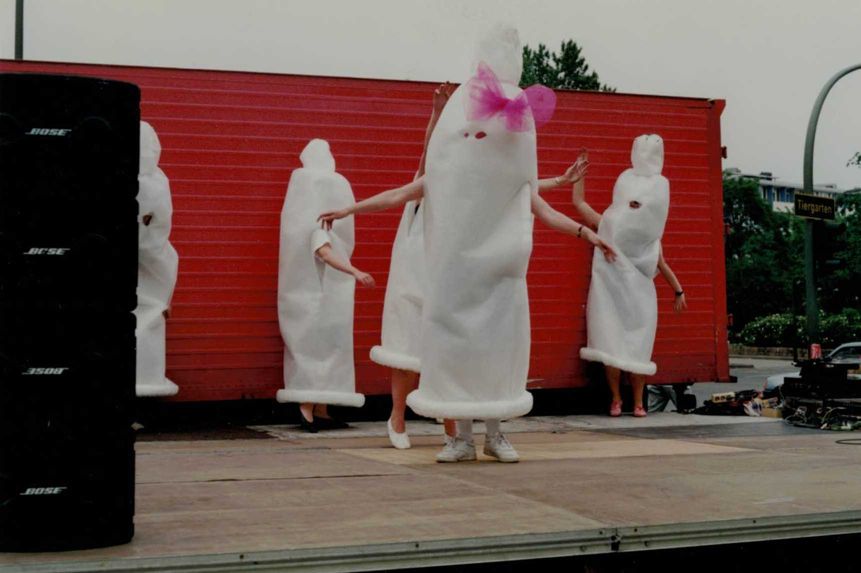 Auf einer Bühne im Freien stehen fünf Personen in langen weißen „Kondomkostümen“, im Hintergrund Straßenschild mit der Aufschrift „Tiergarten“