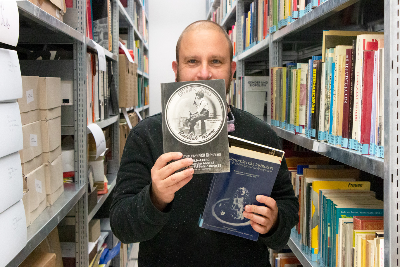 Roman Klarfeld steht zwischen den Archivregalen der FFBIZ-Bibliothek und hält in der rechten Hand das Buch „Biederer Alltag – Radikale Träume, 5. Sommeruniversität für Frauen“. In seiner linken hält er drei weitere Bücher, die die Sommeruniversitäten zum Thema haben..