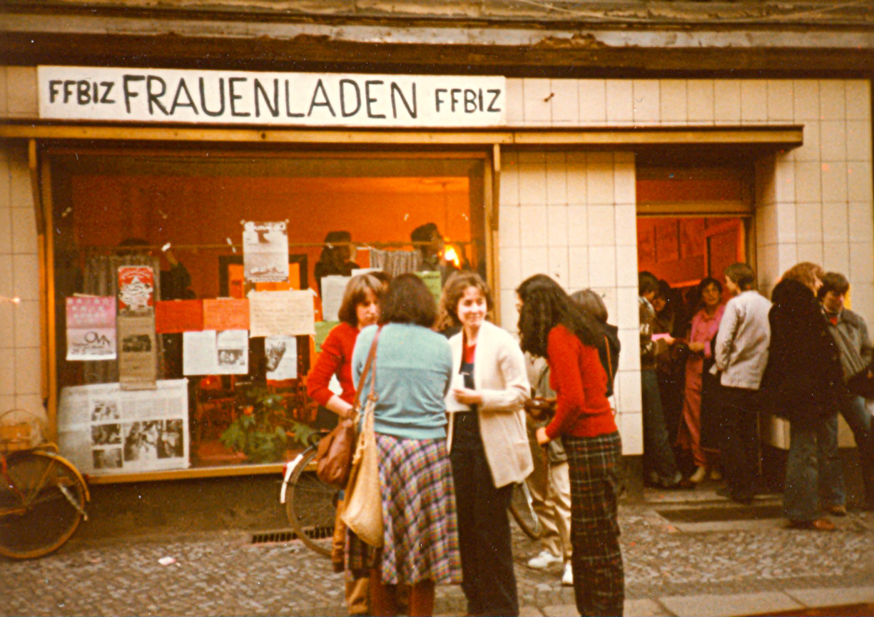 FFBIZ-Mitarbeiterinnen u. a. Ursula Nienhaus haben sich auf dem Bürgersteig vor dem Frauenladen und in den Räumen des FFBIZ versammelt bzw. stehen in Gruppen zusammen in der geöffneten Eingangstür und unterhalten sich, an der großen Fensterfront hängen Plakate mit Veranstaltungshinweisen, über der Fensterscheibe ist das Logo bzw. die Aufschrift „FFBIZ Frauenladen FFBIZ“ sichtbar