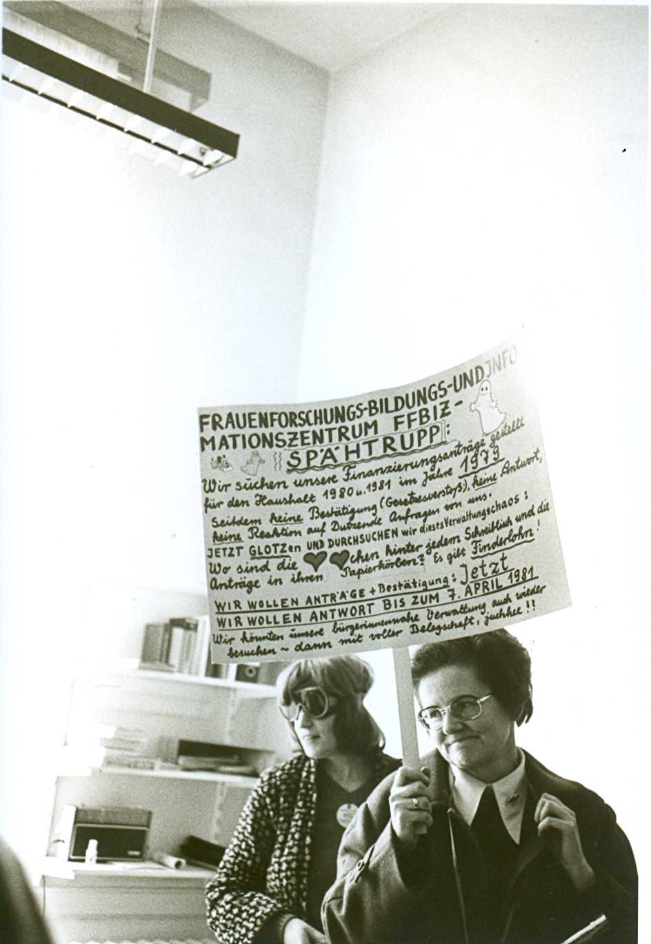 Gisela Vollradt protestiert neben Ursula Nienhaus (v.r.n.l.) in dem Büroraum der Senatsverwaltung Gaus und hält ein Protest-Plakat mit dem folgenden Text hoch: Frauenforschungs-Bildungs und Informationszentrum FFBIZ- Spähtrupp. Wir suchen unsere Finanzierungsanträge gestellt für den Haushalt 1980 u. 1981 im Jahre 1979. Seitdem keine Bestätigung (Gesetzesverstoß), keine Antwort, keine Reaktion auf Dutzende von Anfragen von uns. Jetzt glotzen und durchsuchen wir dieses Verwaltungschaos. Wo sind die Herzchen hinter jedem Schreibtisch und die Anträge in ihren Papierkörben? Es gibt Finderlohn! Wir wollen Anträge und Bestätigungen: Jetzt. Wir wollen Antwort bis zum 7. April 1981. Wir können unsere bürgernahe Verwaltung auch wieder besuchen - dann mit voller Belegschaft. Juchhu!; Ursula Niehaus trägt einen Button an ihrem T-Shirt und eine Schwimmbrille im Gesicht