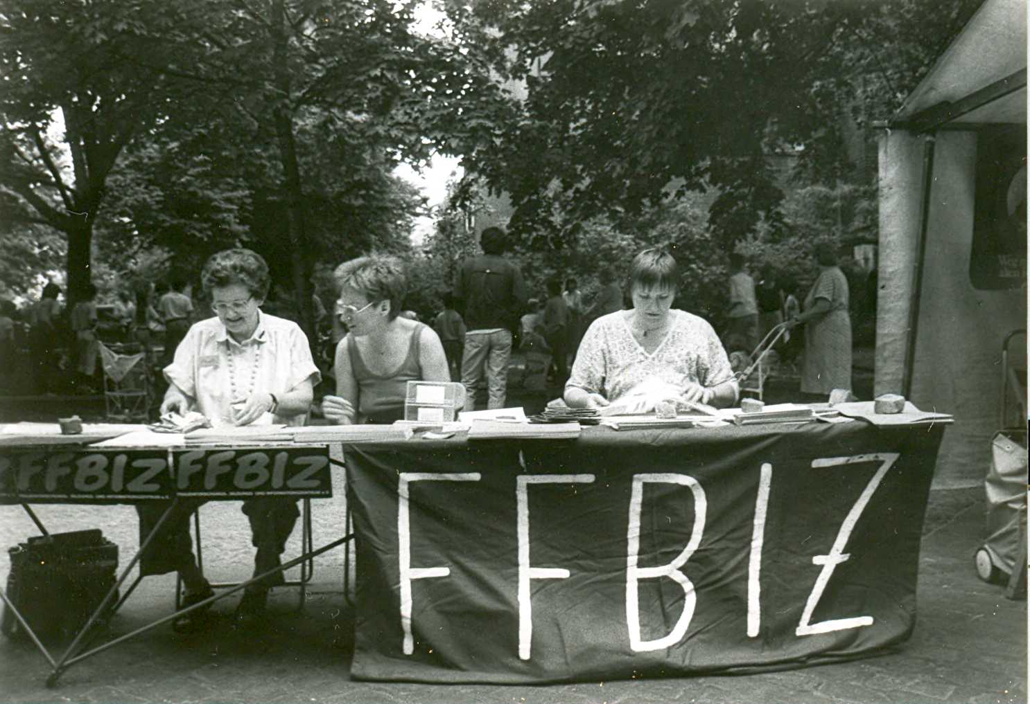 Gisela Vollradt, Ursula Nienhaus und Gabriele Wohlauf (v.l.n.r.) hinter dem mit Broschüren und Infomaterialien bestücktem Infotisch auf dem Karl-August Platz in Charlottenburg; im Hintergrund spielende Kinder und Erwachsene mit Kinderwagen