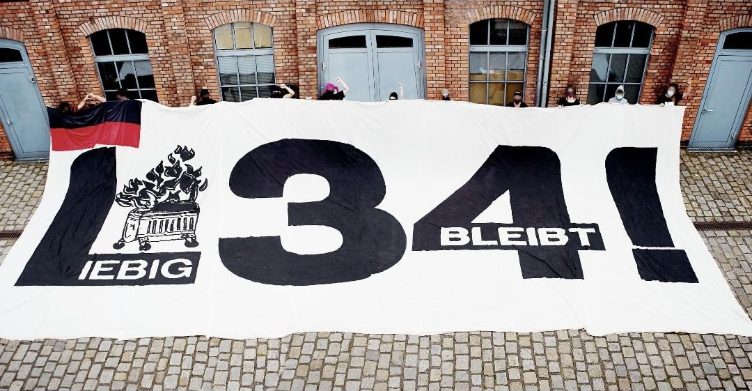 weißes Transparent mit schwarzer und weißer Aufschrift und Graphik eines brennenden Müllcontainer wird von nicht kenntlich gemachten Personen gehalten und liegt ausgebreitet auf dem Kopfsteinpflaster vor einer Gebäudefassade